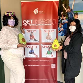 Two women in masks holding avocado toys in front of Heart Association signage